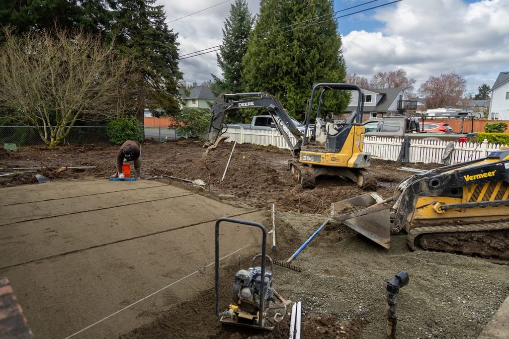 A Father Nature crew working a graded, compacted base on a Tacoma backyard renovation, with a mini excavator and a track loader moving soil under a grey sky