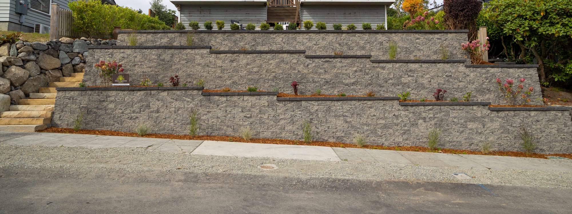 The full street frontage, with three tiers of block retaining wall stepping up the slope and stone steps at the left